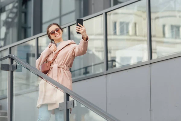 Mulher em vestido rosa tirar selfie em smarthone no fundo urbano cinza ao ar livre — Fotografia de Stock