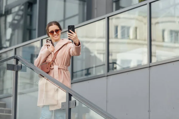 Mulher em vestido rosa tirar selfie em smarthone no fundo urbano cinza ao ar livre — Fotografia de Stock