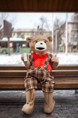 Pelüş bir oyuncak ayı elinde akıllı bir telefonla bankta oturuyor..