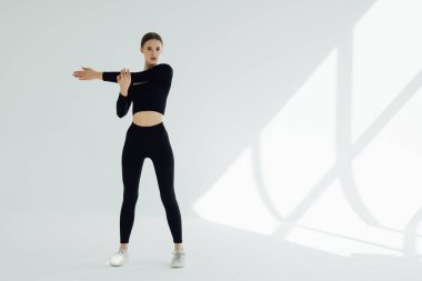 Spor kıyafetli genç beyaz kadın at kuyruğu ile beyaz duvar arkasında duruyor, ellerini uzatıyor.