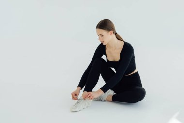 Spor tişörtlü ve spor tozluklu genç bir kadın model yerde oturuyor ve izole edilmiş beyaz bir arka planda ayakkabı bağcıklarını bağlıyor.