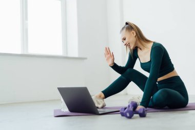Çevrimiçi Egzersiz Maratonu, Uzaktan Eğitim. Mutlu bir milenyum kadını paspasın üzerinde oturup koçla konuşuyor, öğretmeniyle görüntülü görüşme yapıyor, sanal egzersiz için bilgisayar kullanıyor, su içiyor.