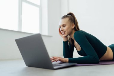 Gülümseme, yoga antrenörüne uyuyor. Mutlu genç spor yoga hocası evde dizüstü bilgisayarla yatıp bilgisayar başında antrenman videosu izliyor. Çevrimiçi fitness öğretim konsepti.