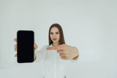 Neşeli sevimli kadın beyaz bir arka plan üzerinde izole smartphone ekranında işaret parmağı. Kameraya bakarak