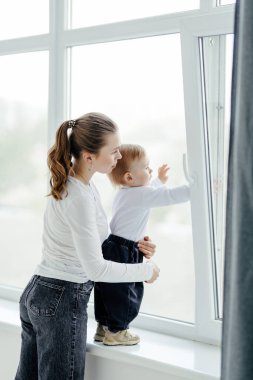 Parlak beyaz bir odada pencerenin yanında oynayan gülümseyen genç bir anne ve oğlunun portresi. Dışarıda güneşli bir gün. Gerçek anne sevgisi