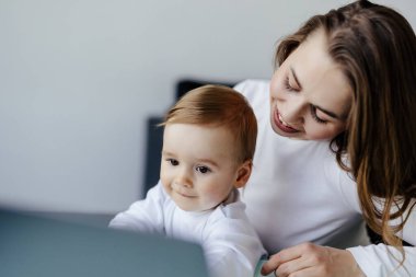 Genç anne ve oğul masada oturuyor ve evde dizüstü bilgisayar kullanıyorlar. Gülümseyen anne e-posta yazarken çocuğuyla birlikte evde çalışıyor..
