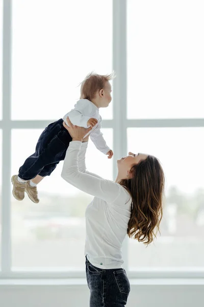 Mom\'s love. Young mom holding toddler son on hands, cuddling with kid boy, standing near window indoors, free space.
