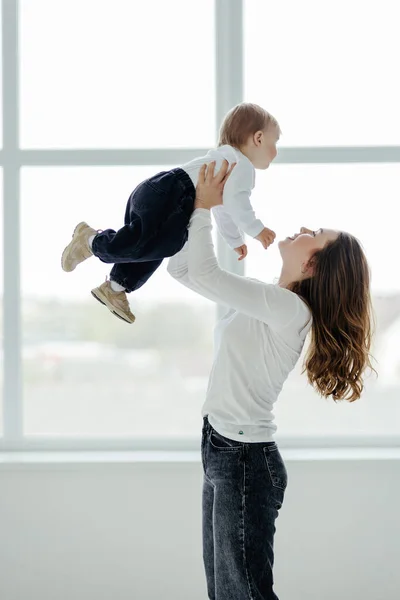 Mom\'s love. Young mom holding toddler son on hands, cuddling with kid boy, standing near window indoors, free space.