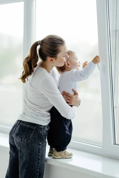 一个笑着的可爱的年轻母亲和她的儿子在一个明亮的白色房间的窗户边玩耍的画像 外面阳光灿烂 真正的母爱 — 图库照片
