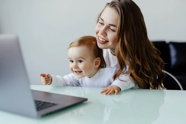 Young beautiful woman multitasking, working from home teaching two year old son to draw. Woman spending quality time with her toddler child.