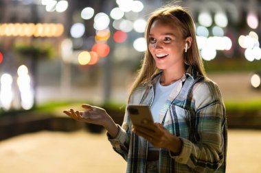 Smiling young woman walking in the city at night with headphones enjoy listening online music from smartphone and looking illuminated city night lights. People and technology concept.