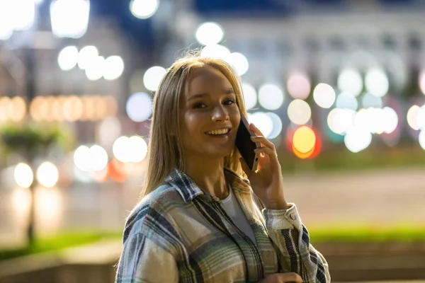 Beautiful young woman talking on smartphone walking through night city street full of neon light. Portrait of smiling female using mobile phone.
