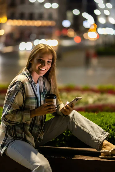 Young Woman Smartphone Sit Bench Night Blurred Lights Building Background Fotos de stock libres de derechos