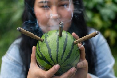 Genç bir kadın elinde karpuzdan yapılmış bir pipo ve boruya tutturulmuş üç marihuana esrarı tutuyor.