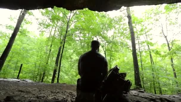 Egy Férfi Egy Barlangban Egy Hátizsákkal Mellette Egy Zöld Erdőt — Stock videók