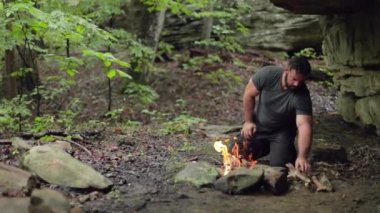 Erkek bir kampçı, yanan bir kamp ateşinin yanında, sığ bir odak noktasıyla odun topluyor..