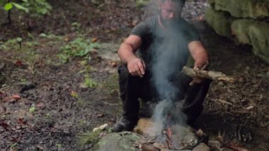 Erkek bir kampçı, yanan bir kamp ateşinin yanında ağır çekimde, yüzeysel bir odak ile oturur..