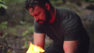 Bir erkek kampçı, yanan bir kamp ateşinin yanında ağır çekimde çalışır..