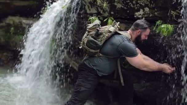 男のハイカーはゆっくりと流れる滝の底の近くに彼の顔を洗う — ストック動画