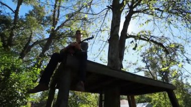 Genç çocuk ağaç platformunda gitar çalıyor, yavaş çekim silueti parlak öğleden sonra güneşiyle arka planda..