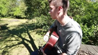 Genç çocuk ağaç platformunda gitar çalıyor. Yavaş çekim, parlak öğleden sonra güneşi altında..
