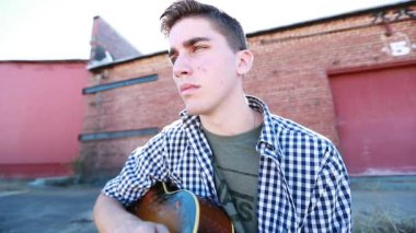 Genç çocuk arka planda eski bir tuğla bina ile çimenlerin yanında yavaş çekimde gitar çalıyor..