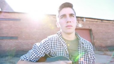 Genç çocuk arka planda eski bir tuğla bina ile çimenlerin yanında yavaş çekimde gitar çalıyor..