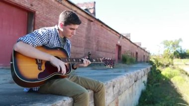 Genç çocuk arka planda eski bir tuğla bina ile çimenlerin yanında yavaş çekimde gitar çalıyor..
