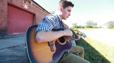 Genç çocuk arka planda eski bir tuğla bina ile çimenlerin yanında yavaş çekimde gitar çalıyor..