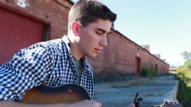Genç çocuk arka planda eski bir tuğla bina ile yavaş çekimde gitar çalar..