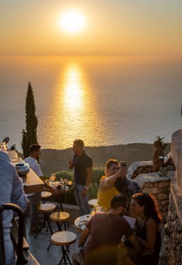 Lefkada adası. Yunanistan - 08.07.2021: İnsanlar ve turistler muhteşem gün batımı manzaralı tepedeki bir restoranda güzel bir yemek yiyorlar.