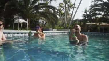 4K 'daki bir kır evinde güneşli bir günde ellerinde bira kutularıyla havuzda kahkahalarla gülen birkaç arkadaş.