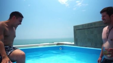 Terasta bir havuzda kadeh kaldıran iki arkadaş deniz ve mavi gökyüzü olan bir sahil evinde viski içiyorlar.