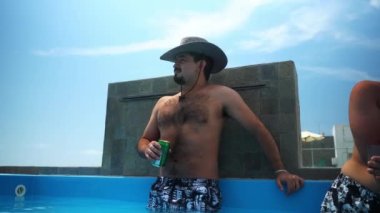 Şapkalı bir adam, elinde birayla bir sahil evindeki terasta havuzdaki soğuk suyun çıkışına şaşırıyor.