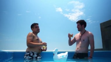 Konuşan ve kadeh kaldıran iki arkadaş, güneşli bir günde güneşli bir terasta, havuzda cam bardaklarla viski içiyorlar.