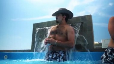 Elinde bira ve şapkası olan bir adam günde 4K ile bir sahil evindeki terasta havuzdaki soğuk su birikintisiyle poz veriyor.