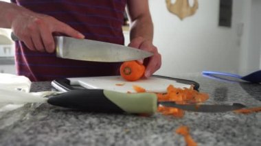 Gündüz vakti beyaz bir adamın elleri mutfakta beyaz bir tahtanın üzerinde havuç keserken görülüyor.