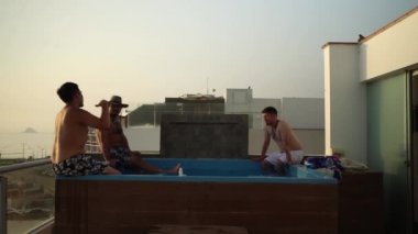 4K 'da gün batımında sahile bakan bir evde yüzme havuzunun kenarında oturan birkaç arkadaş.