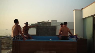 Gün batımında sahile bakan bir evde ayaklarıyla havuz kenarında oturan birkaç arkadaşa bira dağıtıyorlar.