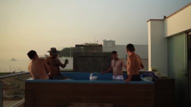 Birkaç arkadaş gün batımında plaj manzaralı bir evdeki yüzme havuzunda elleri ile bira kutularını deviriyor.