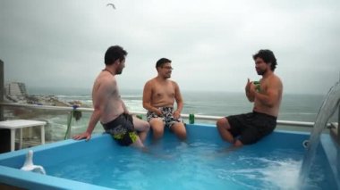 4K bulutlu bir günde, bir sahil evinde, düşen suyla ve okyanus manzaralı bir havuzda bira içen birkaç arkadaşın kamera görüntüsü.