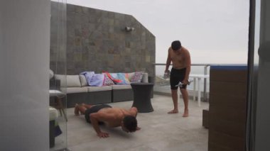 İki arkadaş, gün boyunca arka planda deniz olan bir sahil evinin terasında dambıl ve kalas taşıyarak egzersiz yapıyorlar.