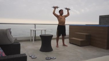 Gün boyunca arka planda deniz olan bir yüzme havuzu olan bir sahil evinin terasında dambıllarla egzersiz yapan bir adamın tam karesi.