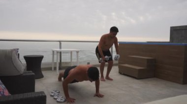 Bulutlu bir günde, arka planda deniz olan bir yüzme havuzu olan bir sahil evinin terasında halter ve kalaslarla egzersiz yapan iki arkadaşın tam karesi.