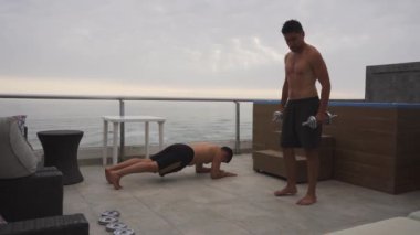 Bulutlu bir günde, arka planda deniz olan bir yüzme havuzu olan bir sahil evinin terasında halter ve kalaslarla egzersiz yapan iki arkadaşın tam karesi.