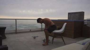 Bir adam bisepsli kıvrımlı egzersizler yapıyor. Deniz kenarında yüzme havuzu olan bir sahil evinin terasında. Bulutlu bir günde martıyı geçiyor.