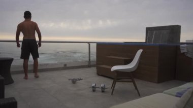 Bir adam havuzu ve şelalesi olan bir sahil evinin terasında dambıllarla egzersiz yaptıktan sonra ufka bakıyor ve bulutlu bir günde arka planda deniz var.