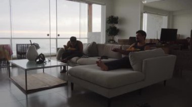 İki arkadaş, bulutlu bir günde plaj evinde kırmızı soslu spagetti ve denizin arka planında tavuk yer ve 4K 'da televizyon izlerler.