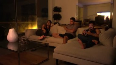 Birkaç arkadaş koltukta oturup televizyon seyrediyor. Deniz manzaralı bir sahil evinin oturma odasında, gece 4K 'de. Bir tanesi cep telefonuna bakıyor.