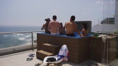 Birkaç arkadaş, güneşli bir günde arka planda deniz olan bir sahil evinin terasında bira ve şarap içerek kadeh kaldırıyor.